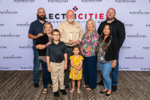A group of eight people, including adults and children, stand together in front of a backdrop that reads 'ELECTRICITIES OF NORTH CAROLINA, INC.' The central figure holds an award plaque.