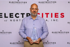 A man is standing in front of a backdrop that reads 'ELECTRICITIES of NORTH CAROLINA, INC.' The person is holding a clear, triangular-shaped award