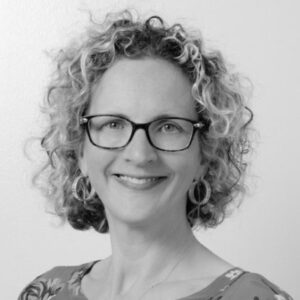 Portrait photograph of a smiling woman with curly hair, glasses, hoop earrings, and a patterned top. 